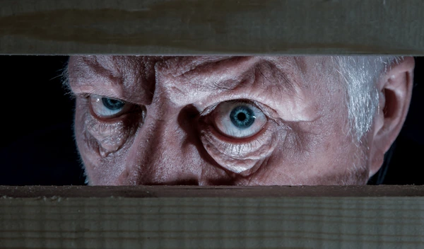 An old man appears to be looking out of the gap between two boards or out of a window. We can only see his eyes, but we can tell his expression is menacing.